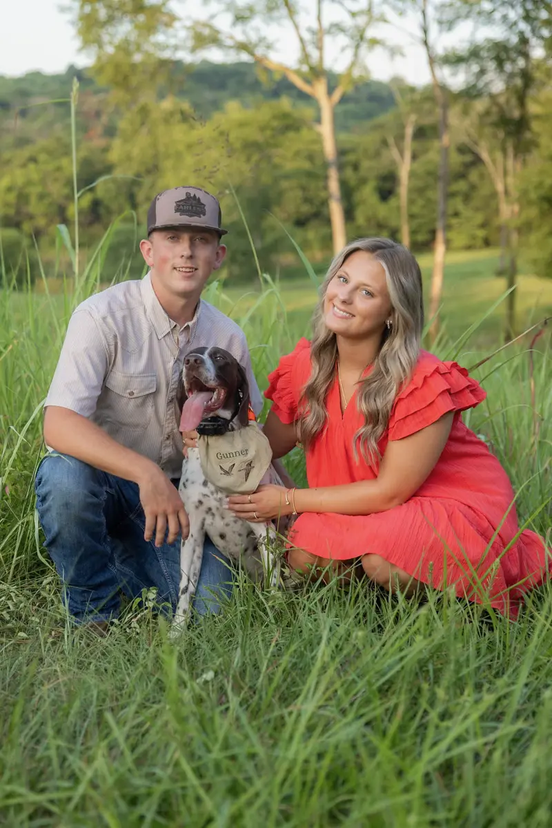 Tyler Farless and Brianna kneeling in tall grass with their bird dog, Gunner